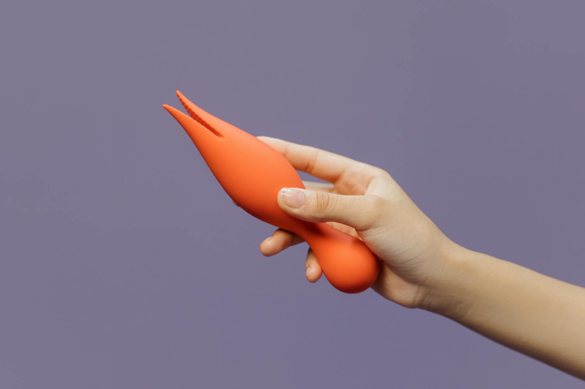 Eine Hand hält einen orangefarbenen Vibrator gegen einen minimalistischen violetten Hintergrund, die moderne Sinnlichkeit zeigt.