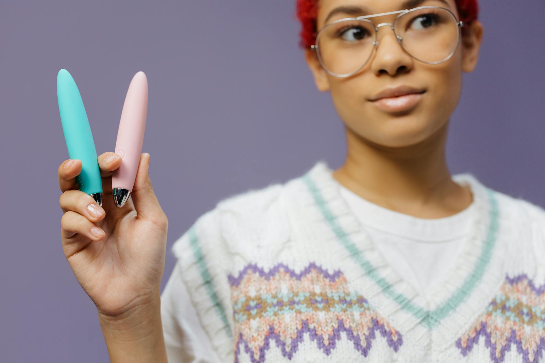 Eine junge Frau mit Brille hält zwei bunte Vibratoren vor einem violetten Hintergrund.